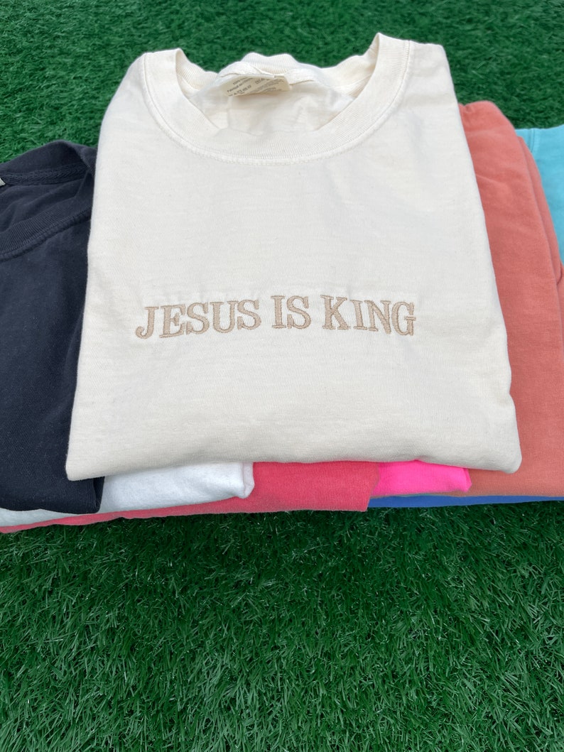May include: A stack of t-shirts in various colors, including black, white, pink, and blue. The top shirt is cream colored with the text "JESUS IS KING" embroidered in brown.