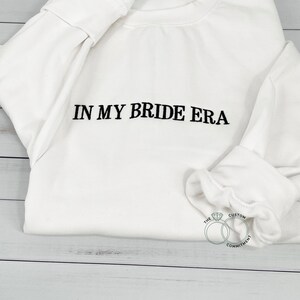 May include: White sweatshirt with the text "IN MY BRIDE ERA" in black embroidery. The bottom of the sweatshirt has a small logo with two interlocking rings and a diamond, with the text "THE CUSTOM COMMITMENT".