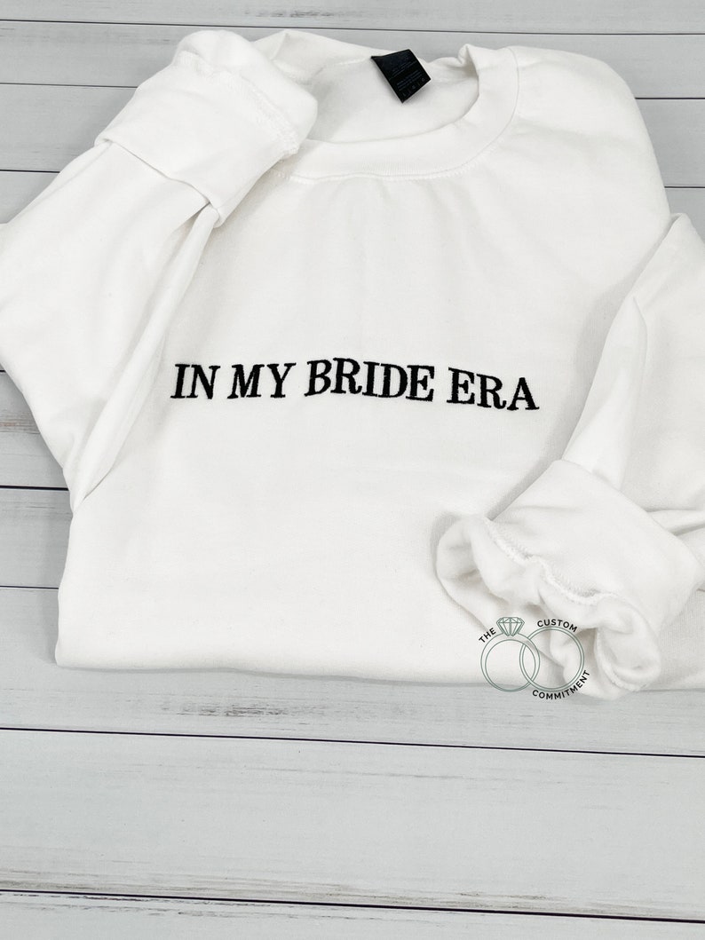 May include: White sweatshirt with the text "IN MY BRIDE ERA" in black embroidery. The bottom of the sweatshirt has a small logo with two interlocking rings and a diamond, with the text "THE CUSTOM COMMITMENT".