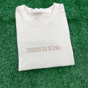 May include: A cream-colored t-shirt with the text "JESUS IS KING" embroidered in brown on the front.