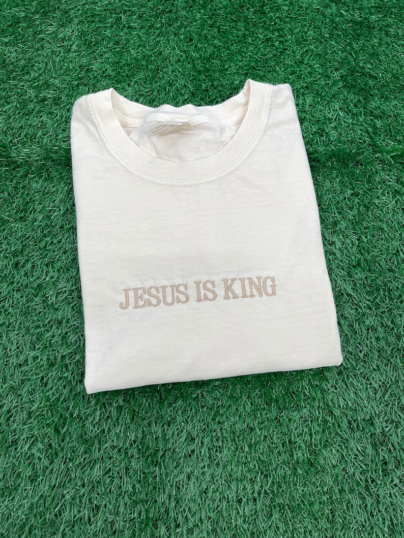 May include: A cream-colored t-shirt with the text "JESUS IS KING" embroidered in brown on the front.
