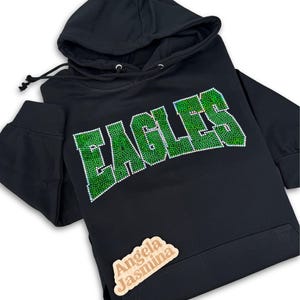 May include: Black hoodie with a green rhinestone "EAGLES" design. The hoodie has a drawstring hood and a small wooden tag with the text "Angela Jasmina" on the bottom right corner.
