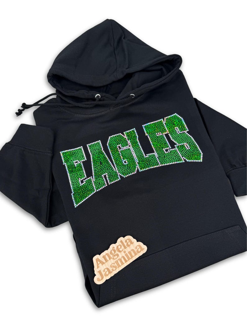May include: Black hoodie with a green rhinestone "EAGLES" design. The hoodie has a drawstring hood and a small wooden tag with the text "Angela Jasmina" on the bottom right corner.