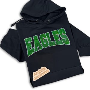 May include: Black hoodie with a green rhinestone "EAGLES" design. The hoodie has a drawstring hood and a kangaroo pocket.