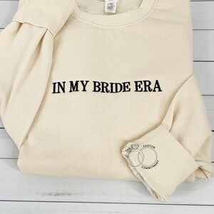 May include: A cream-colored sweatshirt with the text "IN MY BRIDE ERA" embroidered in black. The sweatshirt has a crew neck and long sleeves. The bottom of the sleeve has a small embroidered design of two interlocking rings with the text "THE CUSTOM COMMITMENT".
