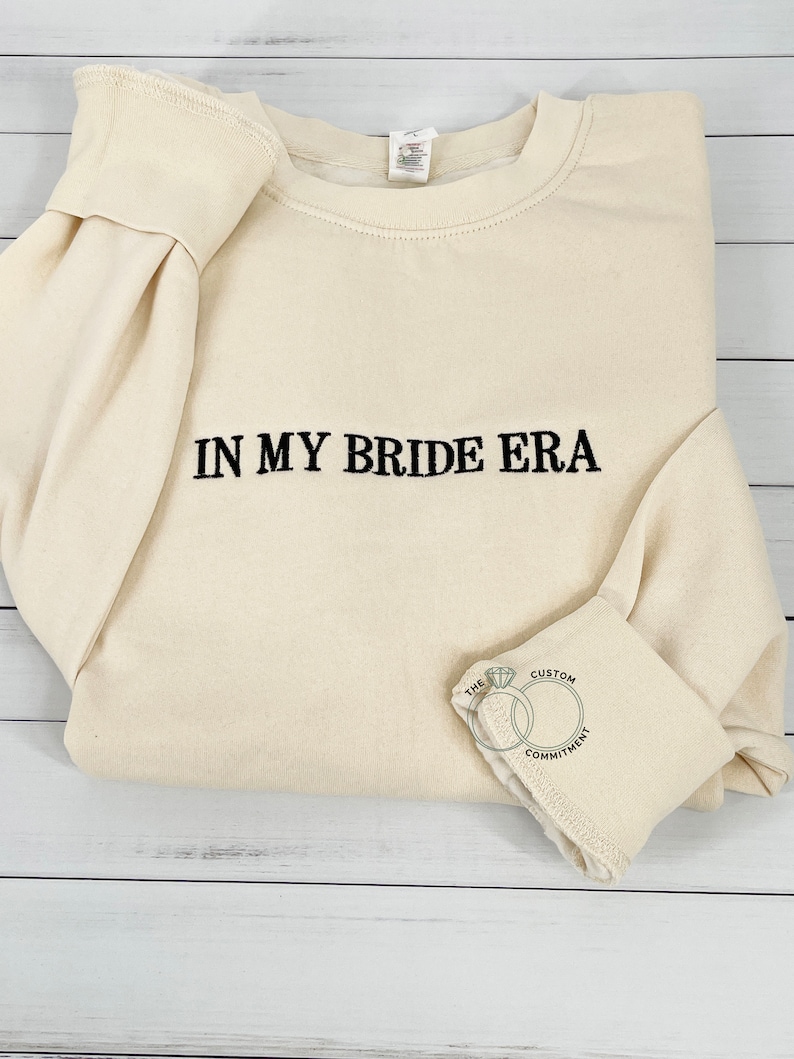 May include: A cream-colored sweatshirt with the text "IN MY BRIDE ERA" embroidered in black. The sweatshirt has a crew neck and long sleeves. The bottom of the sleeve has a small embroidered design of two interlocking rings with the text "THE CUSTOM COMMITMENT".