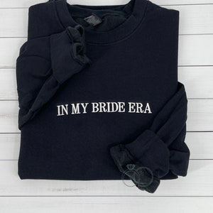 May include: Black long-sleeved sweatshirt with the text "IN MY BRIDE ERA" embroidered in white on the front.