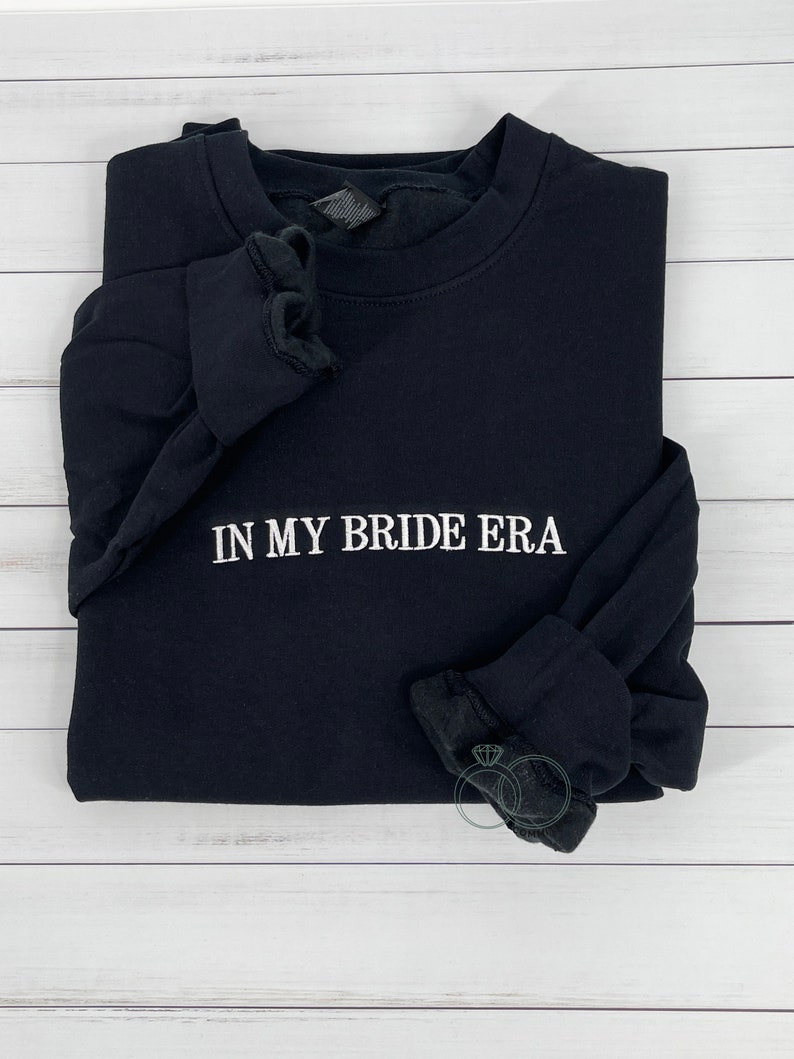 May include: Black long-sleeved sweatshirt with the text "IN MY BRIDE ERA" embroidered in white on the front.