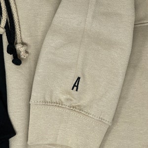 May include: A beige hoodie with a black drawstring and a black embroidered letter 'A' on the sleeve.
