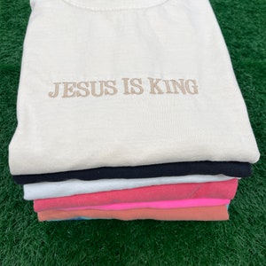 May include: A stack of six t-shirts in various colors, including white, black, pink, and blue. The top t-shirt is cream colored and has the text "JESUS IS KING" embroidered in brown on the front.