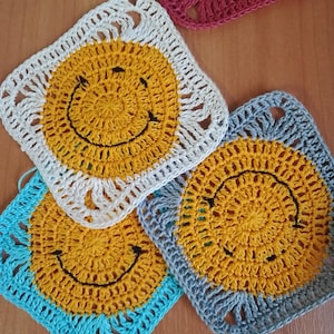 May include: A selection of crocheted squares, each featuring a yellow smiley face with a black stitched mouth and eyes. The squares have borders in white, blue, grey, and maroon. The handmade squares are arranged on a wooden surface.