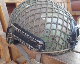 May include: A camouflage helmet with a green mesh net and a black strap. The helmet has a black side rail and a black strap on the back.