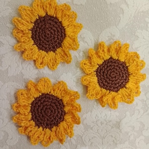 Puede incluir: Tres apliques de girasol tejidos a ganchillo, cada uno con un centro circular marrón oscuro rodeado de pétalos de color amarillo brillante. La textura del hilo es visible en las puntadas circulares de los centros y los pétalos.