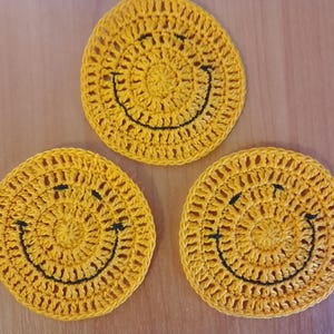 May include: Three yellow crocheted coasters with black smiley faces. The coasters are round and have a textured appearance. The coasters are handmade and are likely used for placing drinks on.