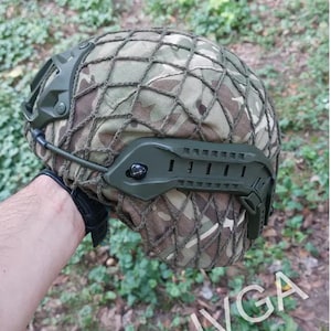 May include: A green camouflage helmet with a mesh cover and a green strap. The helmet has a black strap on the side.