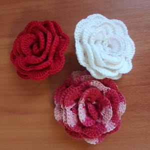 May include: Three handmade crochet flowers in red, white, and a red and pink spotted pattern. The flowers are arranged on a wooden surface. The flowers are likely used for decoration or as embellishments.