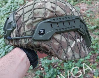 May include: A green camouflage helmet with a mesh cover and a green side rail system. The helmet is being held by a hand wearing a black watch.