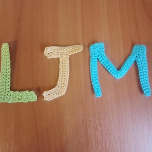 Puede incluir: Letras de ganchillo L, J y M, en verde claro, amarillo y azul turquesa, respectivamente, dispuestas sobre una superficie de madera. Las letras hechas a mano probablemente forman parte de un proyecto de artesanía personalizado o un artículo decorativo.