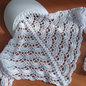 Puede incluir: Un chal blanco de ganchillo con un delicado patrón calado. El chal está drapeado sobre un objeto blanco de forma ovalada, posiblemente una horma de sombrero. El intrincado diseño presenta motivos circulares repetidos, creando una textura de encaje.