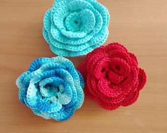 May include: Three handmade crochet flowers in shades of blue and red. The flowers are arranged on a light brown wooden surface. The flowers are in the shape of roses.