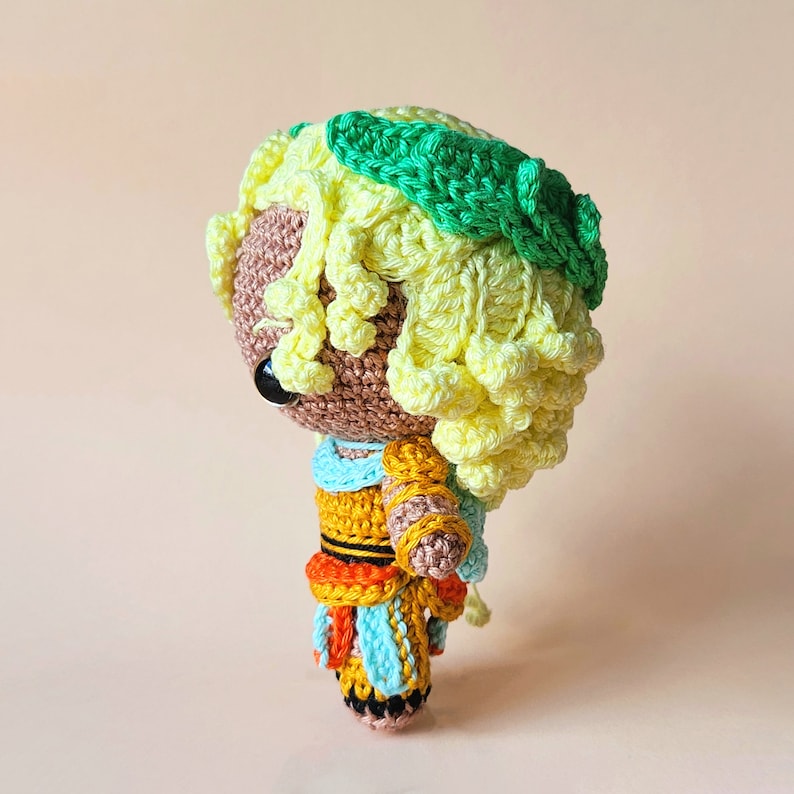 Peut inclure: Une poup&eacute;e en crochet avec des cheveux jaunes, une couronne verte et une robe color&eacute;e. La poup&eacute;e a des yeux bruns et un visage brun clair.