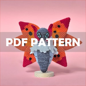 May include: A crochet moth figurine featuring large orange and red wings with black spots, a dark gray head with light blue eyes, and a white fluffy collar. The moth has a gray body and stands on a small circular wooden base. Text reads PDF PATTERN.