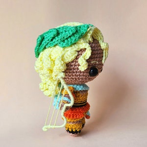 Peut inclure: Une poup&eacute;e en crochet avec des cheveux jaunes, une couronne verte et une robe color&eacute;e. La poup&eacute;e tient une petite harpe en crochet blanche.