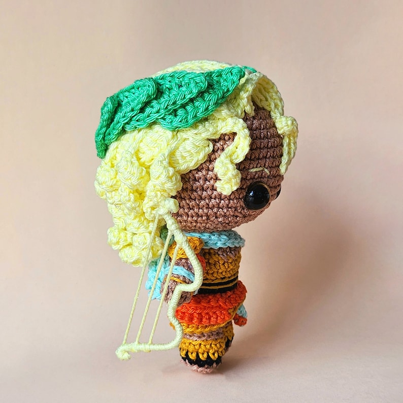 Peut inclure: Une poup&eacute;e en crochet avec des cheveux jaunes, une couronne verte et une robe color&eacute;e. La poup&eacute;e tient une petite harpe en crochet blanche.