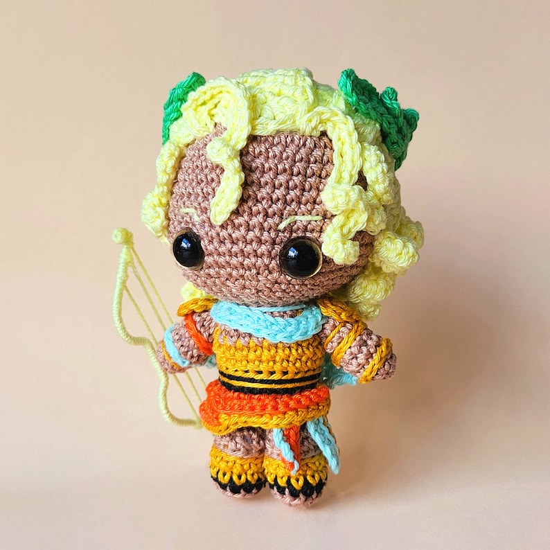 Peut inclure: Une poup&eacute;e amigurumi en crochet avec des cheveux jaunes, des feuilles vertes et une tenue bleue et orange. La poup&eacute;e tient un arc et une fl&egrave;che jaunes.