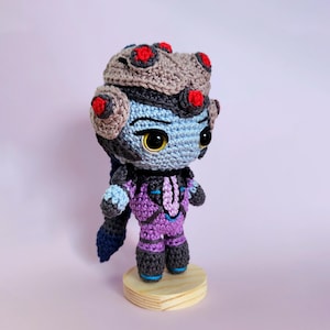 May include: A handmade crochet doll featuring a blue-skinned character with a gray and red headpiece. The doll wears a purple and black jumpsuit and stands on a small wooden base. The doll has large, expressive eyes and is made with detailed stitching.