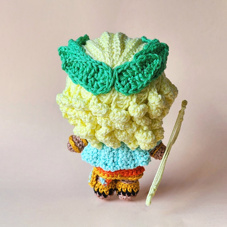 Peut inclure: Une poup&eacute;e en crochet avec des cheveux jaunes et verts, portant une robe bleue et orange et tenant un arc vert et blanc.