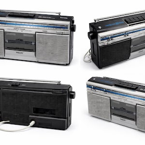 Puede incluir: Un radiocasete Philips vintage en plata y negro, con reproductor de casetes, dial de radio y antena retráctil. El radiocasete tiene un asa y rejillas de altavoz. El logotipo de Philips es visible.
