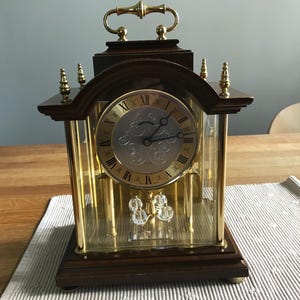 May include: A mantel clock with a dark wood base and top, featuring a gold-tone metal frame and glass panels. The circular clock face has black Roman numerals and ornate silver-tone detailing. A gold handle sits on top, and a pendulum with four clear spheres hangs inside.
