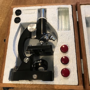 Op de afbeelding: Een vintage zwarte microscoop met zilveren accenten, gehuisvest in een houten koffer met witte schuimvulling. De koffer bevat ook rode ronde componenten en glazen dia's. Een wetenschappelijk instrument.