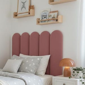 May include: A bedroom scene with a bed featuring a pink headboard, grey bedding with white star patterns, and a cream blanket. A small white bedside table holds a lamp and a plant. The room has wooden shelves and a round rug.