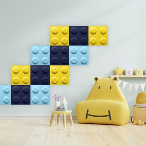 K&ouml;nnte beinhalten: Eine gelbe und blaue schallabsorbierende Wandverkleidung aus Filz, geformt wie ein Stapel Bausteine. Die Verkleidung ist an einer hellblauen Wand in einem Kinderzimmer angebracht. Ein gelber Sessel mit einem l&auml;chelnden Gesicht steht vor der Verkleidung.