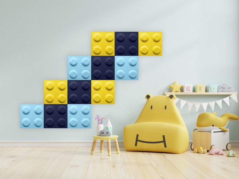 K&ouml;nnte beinhalten: Eine gelbe und blaue schallabsorbierende Wandverkleidung aus Filz, geformt wie ein Stapel Bausteine. Die Verkleidung ist an einer hellblauen Wand in einem Kinderzimmer angebracht. Ein gelber Sessel mit einem l&auml;chelnden Gesicht steht vor der Verkleidung.