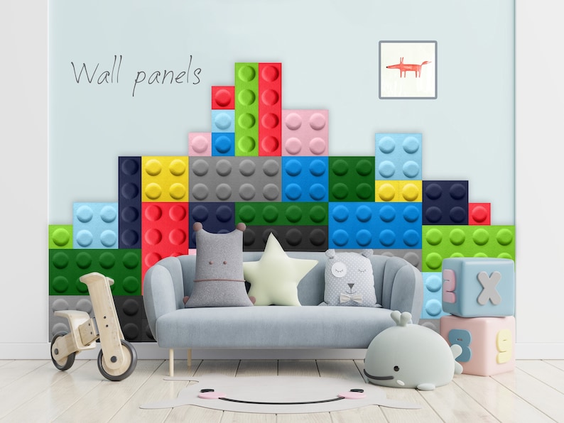 K&ouml;nnte beinhalten: Eine Wand, die mit bunten, weichen, schallabsorbierenden Wandpaneelen dekoriert ist, die gro&szlig;en Lego-Steinen &auml;hneln. Die Paneele sind in einem Muster angeordnet und in den Farben Rot, Blau, Gr&uuml;n, Gelb und Grau gehalten. Ein hellblaues Sofa mit grauen Kissen steht vor der Wand. Ein h&ouml;lzernes Spielzeug-Dreirad befindet sich auf der linken Seite des Bildes. Auf der rechten Seite des Bildes befinden sich au&szlig;erdem zwei kleine, bunte Spielzeugbl&ouml;cke.