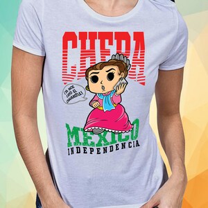 May include: White t-shirt with a red graphic of a cartoon woman holding a phone. The graphic says "CHEDA" and "MEXICO INDEPENDENCIA". The woman is wearing a pink dress and a blue shawl. The text in a speech bubble says "Ya nos cay&oacute; el chahuistle!"