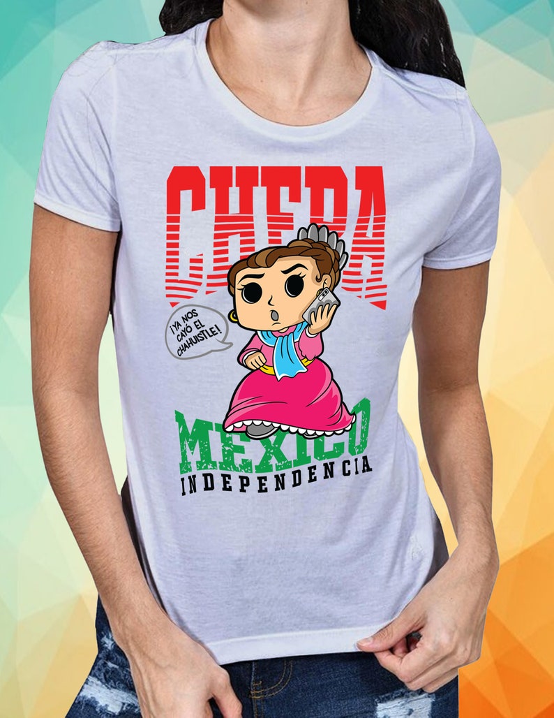 May include: White t-shirt with a red graphic of a cartoon woman holding a phone. The graphic says "CHEDA" and "MEXICO INDEPENDENCIA". The woman is wearing a pink dress and a blue shawl. The text in a speech bubble says "Ya nos cay&oacute; el chahuistle!"