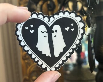 May include: Black and white heart-shaped sticker with two ghosts inside. The ghosts are facing each other and have small hearts around them.