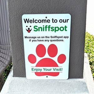 May include: A green and white sign with the text "Welcome to our Sniffspot" and a cartoon dog face. The sign also has a red paw print and the text "Message us on the Sniffspot app if you have any questions. Enjoy Your Visit!"