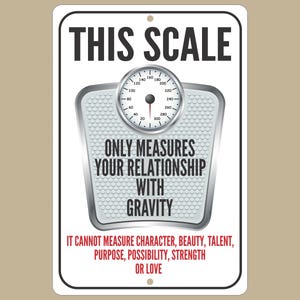 May include: White sign with black text that reads "THIS SCALE ONLY MEASURES YOUR RELATIONSHIP WITH GRAVITY IT CANNOT MEASURE CHARACTER, BEAUTY, TALENT, PURPOSE, POSSIBILITY, STRENGTH OR LOVE". A silver scale with a white face is in the center of the sign.
