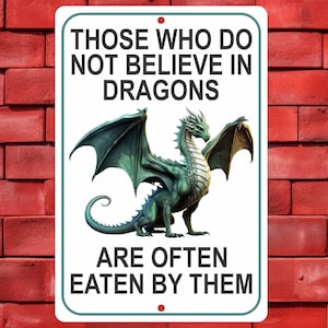 May include: A rectangular white metal sign with rounded corners and two mounting holes. The text reads: THOSE WHO DO NOT BELIEVE IN DRAGONS ARE OFTEN EATEN BY THEM. A green dragon with large wings and a long tail is centered on the sign.