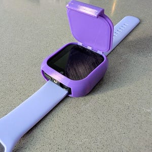 Puede incluir: Una funda de reloj inteligente morada con una tapa con bisagras, abierta para revelar la esfera del reloj. La funda está sujeta a una correa de reloj morada clara. El reloj y la correa están sobre una superficie gris.
