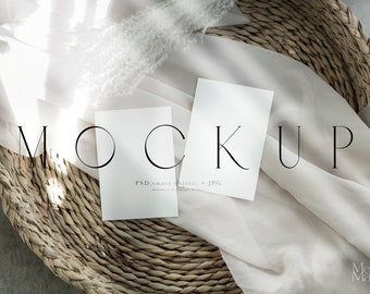 May include: Two blank white cards on a woven basket with a white fabric draped over it. The text "MOCKUP" is written in black on the fabric. The text "PSD (smart object) + JPG" is written in black on one of the cards.