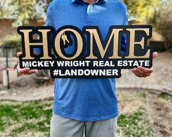 May include: A black and gold wooden sign with the word "HOME" in large gold letters. The sign also includes the text "Mickey Wright Real Estate #LANDOWNER".