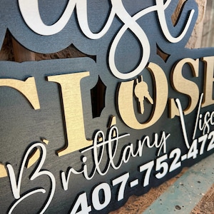 Pu&ograve; includere: Un'insegna in legno a strati con base nera e lettere in rilievo bianche e dorate. Il testo recita "JUST CLOSED" con un'icona a forma di chiave nella lettera O, seguito dal nome "Brittany Visc" e dal numero "407-752-472".