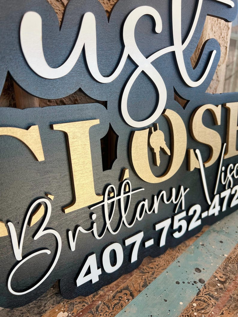 Pu&ograve; includere: Un'insegna in legno a strati con base nera e lettere in rilievo bianche e dorate. Il testo recita "JUST CLOSED" con un'icona a forma di chiave nella lettera O, seguito dal nome "Brittany Visc" e dal numero "407-752-472".