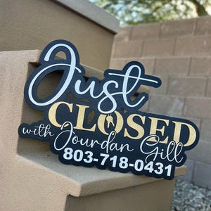 Pu&ograve; includere: Un cartello nero con scritte bianche e dorate che recita Just CLOSED with Jourdan Gill 803-718-0431. La parola Just &egrave; in carattere corsivo bianco, CLOSED in lettere dorate con un'icona a forma di chiave e il resto &egrave; in bianco.